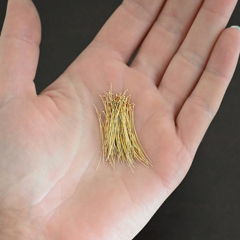 May include: A pile of gold-colored head pins, approximately 1 inch long, are displayed on a person's open palm.