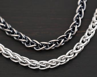 May include: Two silver chains, one with a black oxidized finish, are braided together. The chains are on a dark brown wooden surface.