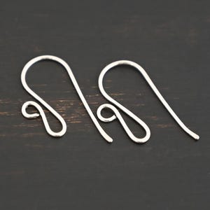 May include: Two silver earring hooks, also known as French hooks, are shown on a dark wood surface. The hooks are made of metal and have a smooth, polished finish.