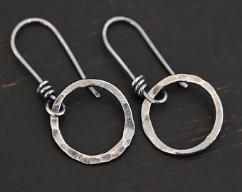 May include: A pair of silver-toned hoop earrings with a hammered texture. The earrings have a simple, open circle design and long, curved ear wires. The background is a dark, wooden surface.