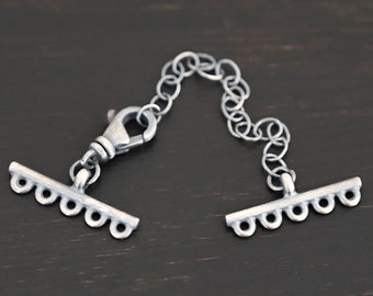 May include: A silver-toned necklace extender with a lobster clasp and decorative bar ends. The extender features a delicate chain and is designed to add length to necklaces or bracelets. The metal has a matte finish.