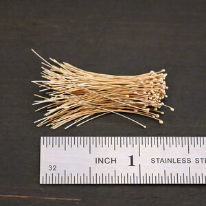 May include: A pile of gold-colored wire with small round beads at the end of each wire. The wire is about 1 inch long. A ruler is in the background.