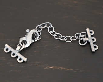 May include: Silver-toned metal clasp with a chain and a toggle closure. The clasp has a hook and loop design. The toggle closure has three loops.