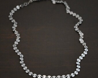 May include: A silver necklace with a chain made of small, round discs. The necklace is on a dark brown surface.