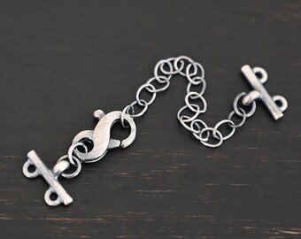 May include: A silver-toned jewelry extender with a lobster clasp, a chain of small oval links, and a T-bar clasp. The extender is designed to add length to necklaces or bracelets. The metal has a slightly oxidized finish.