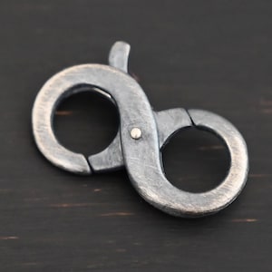 May include: A silver-toned metal clasp with an infinity shape. The clasp has a brushed finish and a small, round, gold-toned accent.