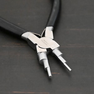 May include: Close-up of a silver-toned jewelry pliers with black handles. The pliers have three prongs for making different sized loops. The pliers are labeled "MAKERSTAR STAINLESS".
