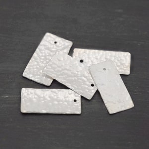 Puede incluir: Cinco etiquetas rectangulares de metal plateado con textura martillada y un agujero en cada esquina. Las etiquetas están apiladas sobre una superficie gris oscuro.
