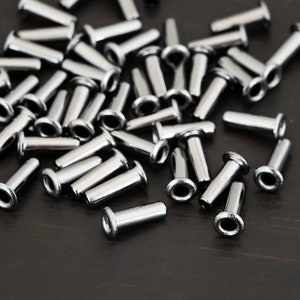May include: A pile of silver metal eyelets, also known as grommets, for crafts and DIY projects. The eyelets have a smooth, shiny finish and are scattered on a dark brown surface.