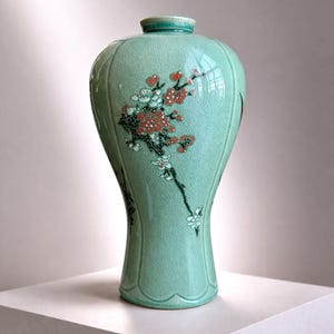 May include: A celadon green ceramic vase with a floral design. The vase features a crackle glaze and a design of red and white flowers on a branch. The vase is displayed on a white surface.