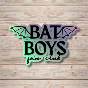 May include: A sticker featuring the text BAT BOYS fan club in black lettering. The design includes stylized bat wings extending from the sides of the word BAT. The background of the sticker has a gradient transitioning from light green to purple.