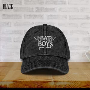 Może przedstawiać: Czarna czapka bejsbolówka z białym haftem "BAT BOYS from club". Czapka ma zakrzywiony daszek i postarzany, vintage wygląd. Słowo "BLACK" jest wydrukowane w lewym górnym rogu.