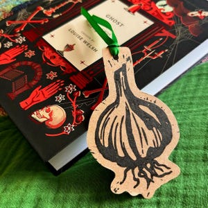 May include: A black and white illustration of a garlic bulb on a brown paper bookmark. The bookmark is tied with a green ribbon and is resting on top of a black book with red and white illustrations. The book title is "Ghost" by Louise Welsh.