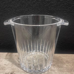 May include: A clear, faceted ice bucket with two handles. The bucket has a flared shape and vertical etched lines. The bucket is made of a clear material and is sitting on a wooden surface.