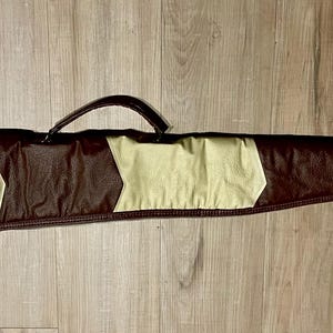 May include: A brown and beige padded rifle case with a handle. The case has a rectangular shape with a zipper closure and a beige chevron pattern. The case is designed to protect a long, slender object.