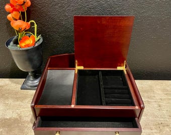 May include: A wooden jewelry box with a dark cherry finish. The box has an open lid, revealing a black velvet interior with compartments. A drawer is visible at the bottom, and gold-colored knobs are on the drawer. A decorative vase with orange flowers is in the background.