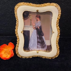 Peut inclure: Un cadre photo rectangulaire avec une bordure dorée ornée et un cadre intérieur crème lisse. Le cadre contient une impression d'une personne en robe claire avec un châle sombre près d'une cheminée. Une fleur orange est placée à côté.
