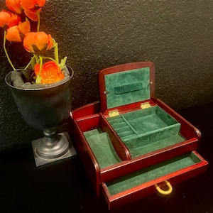 Puede incluir: Un joyero de madera con un acabado marrón rojizo intenso, con un interior de terciopelo verde. La caja está abierta, revelando compartimentos para guardar. Un pequeño jarrón con flores naranjas está a la izquierda.