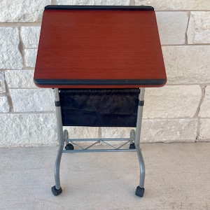 May include: A small, mobile, wooden desk with a reddish-brown surface and black trim. The desk has a black fabric storage pouch and a silver metal frame with wheels. The desk is designed for portability and convenience.