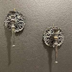 May include: Two ornate wall sconces with a vintage aesthetic. Each sconce features a circular, filigree design surrounding a central candle holder and a long, tapered stem. The sconces are finished in a metallic bronze and silver color scheme.