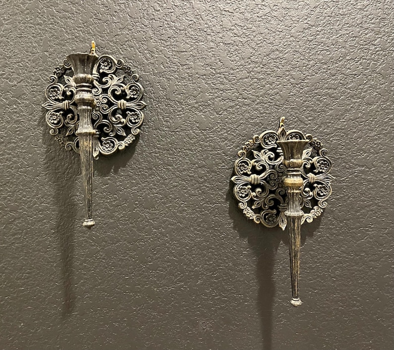 May include: Two ornate wall sconces with a vintage aesthetic. Each sconce features a circular, filigree design surrounding a central candle holder and a long, tapered stem. The sconces are finished in a metallic bronze and silver color scheme.