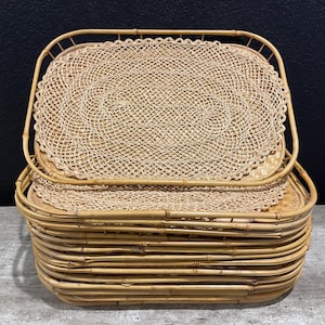May include: A stack of woven bamboo trays and placemats. The trays are rectangular with rounded corners and handles. The placemats have a circular woven design. The items are a natural tan color.