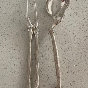 Op de afbeelding: Zilveren serveervork en soeplepel set. De vork heeft twee tanden en de lepel heeft een afgeronde kom. Beide stukken hebben een uniek, getextureerd ontwerp, met een botachtig uiterlijk. De set is gemaakt van een glanzend, reflecterend metaal.