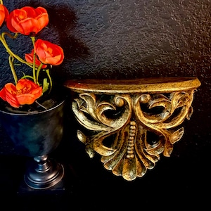 May include: A decorative gold-coloured wall shelf with detailed scrollwork and a semi-circular top. The shelf is mounted on a black wall. A black vase with red flowers is to the left of the shelf. The shelf is made of a dark gold material.