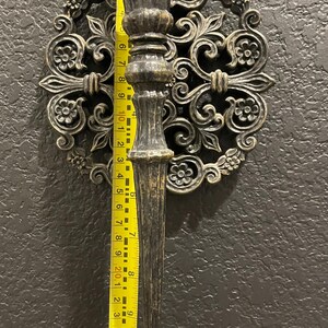 May include: Ornate, antique-style wall sconce with a dark bronze finish. The sconce features a decorative circular backplate with floral and scrollwork details. The sconce itself is approximately 10 inches long, as measured by a yellow ruler.