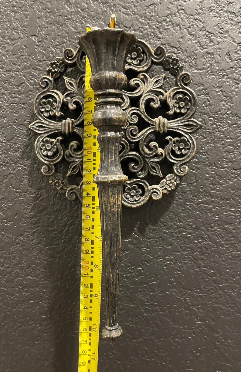 May include: Ornate, antique-style wall sconce with a dark bronze finish. The sconce features a decorative circular backplate with floral and scrollwork details. The sconce itself is approximately 10 inches long, as measured by a yellow ruler.