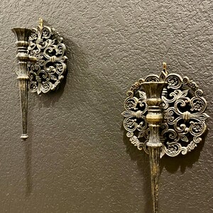 May include: Two ornate, antique-style wall sconces with a bronze finish are mounted on a textured, dark gray wall. Each sconce features a decorative, circular backplate with swirling floral patterns and a slender, candle-holding arm.