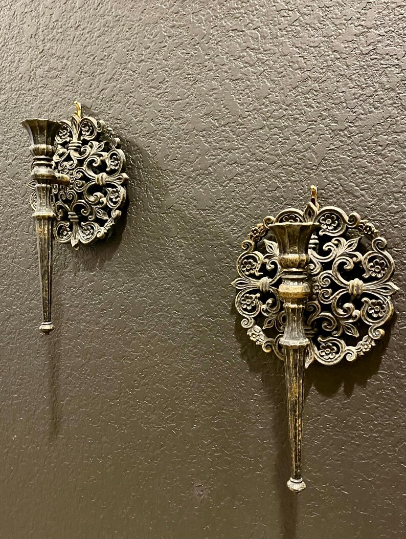 May include: Two ornate, antique-style wall sconces with a bronze finish are mounted on a textured, dark gray wall. Each sconce features a decorative, circular backplate with swirling floral patterns and a slender, candle-holding arm.