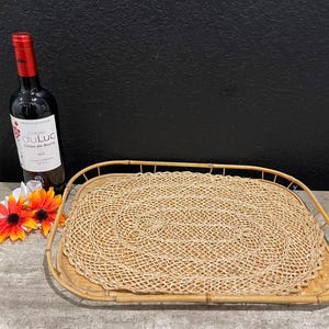 Può includere: Un vassoio rettangolare intrecciato con un tappetino intrecciato circolare all'interno. A sinistra del vassoio ci sono una bottiglia di vino rosso e fiori arancioni e gialli. La bottiglia di vino ha un tappo rosso e un'etichetta bianca.