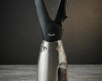 May include: A Chef'n branded spice grinder with a black V-shaped handle and a stainless steel and clear plastic body. The base contains black peppercorns visible through the transparent section. The device features a sleek, modern design.