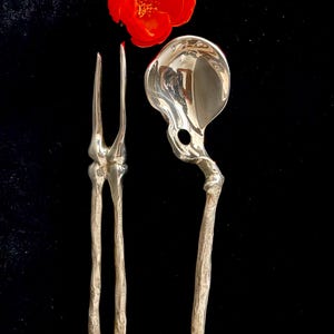 May include: A silver-toned metal fork and spoon set with handles and heads shaped like human skeletal bones, specifically a femur and pelvic structure, accompanied by a single vibrant red flower positioned at the top center of the utensils.
