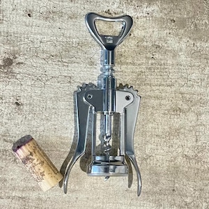 May include: A chrome wine opener with a corkscrew and lever design, next to a wine cork. The opener has a bottle cap opener at the top. The cork is tan with a purple top and printed text.
