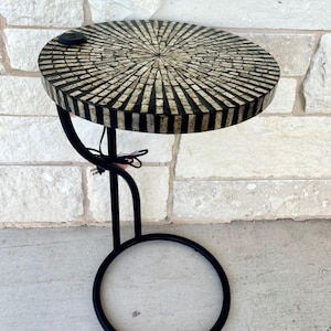 May include: A round side table featuring a black metal frame with a circular base and a curved support arm. The tabletop is inlaid with a radial pattern of rectangular light and dark tiles and includes a small black electrical power outlet port.