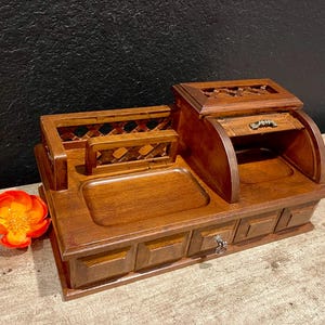 May include: A brown wooden desk organizer featuring a rectangular tray, a lattice-style back panel, and a raised compartment with a small drawer. The base includes three additional drawers with metal pulls, and a bright orange flower sits nearby.
