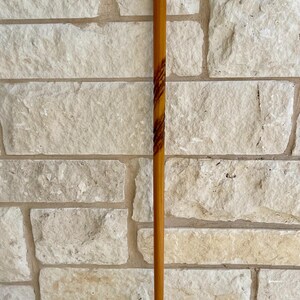 May include: A wooden walking cane with a curved handle and a dark brown accent. The cane is a light brown color with a dark brown spiral design. The tip of the cane has a black rubber cap.