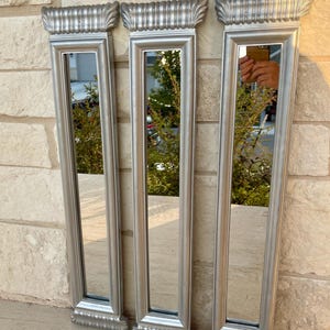 Puede incluir: Tres espejos con marco plateado con un diseño de columna clásica. Cada espejo tiene forma rectangular con elementos decorativos superiores e inferiores. Los espejos se exhiben contra una pared de piedra texturizada, reflejando el entorno.
