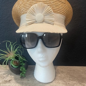 Puede incluir: Un sombrero de paja con una cinta color crema y un adorno decorativo en forma de abanico. El sombrero se combina con gafas de sol negras sobre una cabeza de maniquí. Una pequeña planta en maceta está en el fondo.