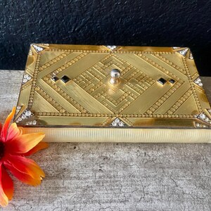 Puede incluir: Una caja rectangular dorada con un diseño Art Déco. La tapa presenta un patrón geométrico con detalles de cristal negro y transparente. Un pequeño pomo plateado sirve de asa. La caja se asienta sobre una base de color crema.
