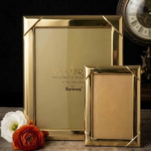May include: Two rectangular gold-toned metal picture frames with mitered corners. The larger frame features text reading SOLID BRASS hand polished & lacquered 8 x 10 (20cm x 25cm) by Bowon. A white and an orange flower are placed in front of the frames.