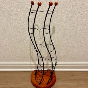 Peut inclure: Un casier à vin en métal noir avec une base en bois. Le rack a un design incurvé avec quatre sections pour contenir des bouteilles. Le haut du rack a quatre sphères orange-brun. La base est de forme semi-circulaire brun rougeâtre.