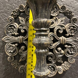 May include: A dark bronze candlestick holder with a decorative floral design. The candlestick is approximately 10 inches tall. The background features a matching wall decor piece with an ornate, symmetrical pattern.