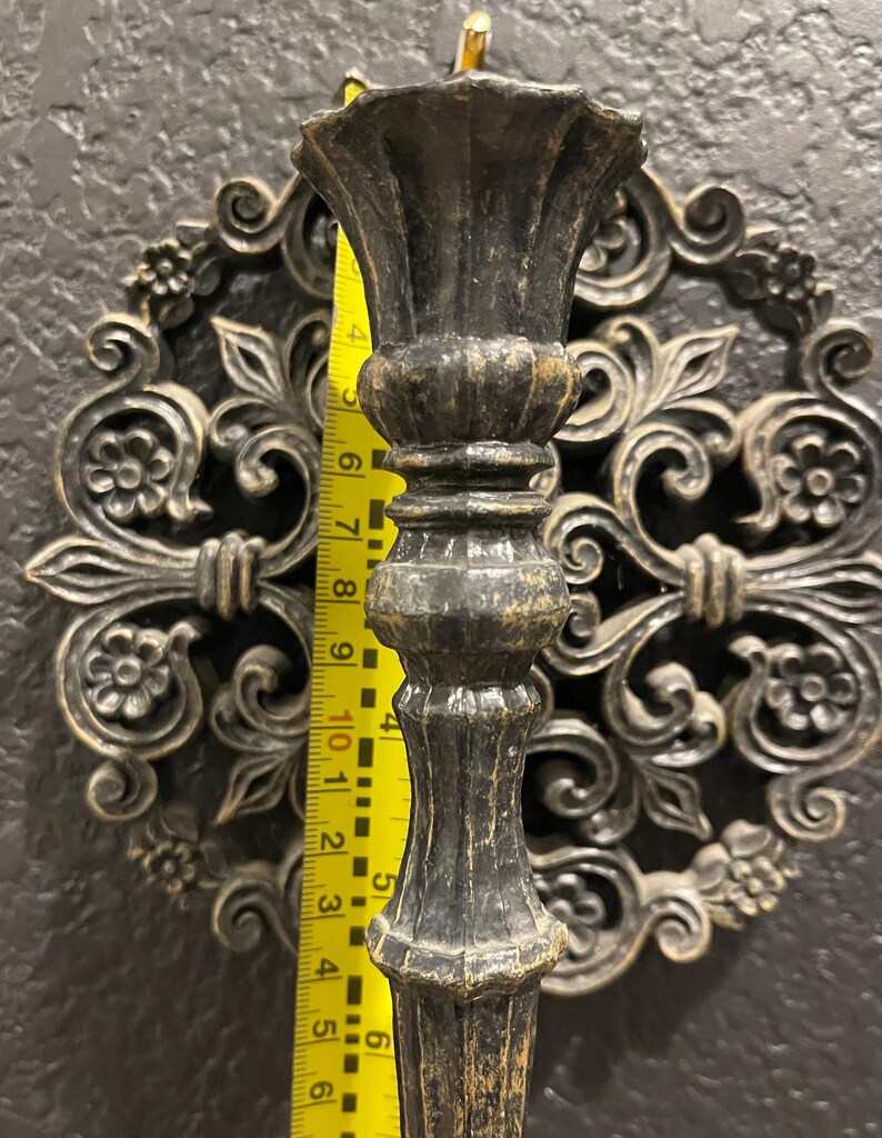 May include: A dark bronze candlestick holder with a decorative floral design. The candlestick is approximately 10 inches tall. The background features a matching wall decor piece with an ornate, symmetrical pattern.