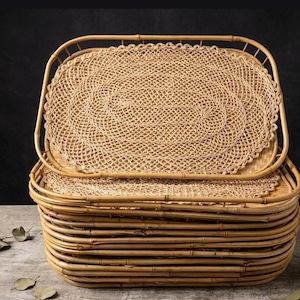 May include: A stack of rectangular bamboo trays with rounded corners, each featuring a light brown woven bamboo base and a natural bamboo frame. The top tray is topped with an oval-shaped, light tan open-weave fiber placemat.