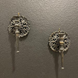 May include: Two ornate wall sconces with a silver finish are mounted on a dark gray textured wall. Each sconce features a circular, floral-patterned backplate and a long, tapered candle holder. The sconces are designed to hold candles and add a decorative touch.