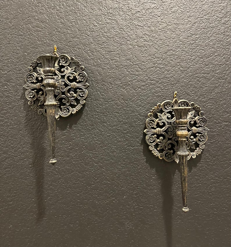 May include: Two ornate wall sconces with a silver finish are mounted on a dark gray textured wall. Each sconce features a circular, floral-patterned backplate and a long, tapered candle holder. The sconces are designed to hold candles and add a decorative touch.