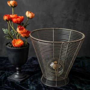 May include: A conical wire basket made of thin silver-toned metal coils with a wide open top and a narrow base. Beside it sits a matte black pedestal vase containing a cluster of vibrant orange artificial flowers with green leaves.